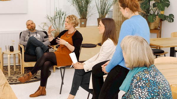 Fünf Personen in einem hellen Raum bei einem Workshop im Gespräch, eine Frau spricht und gestikuliert.