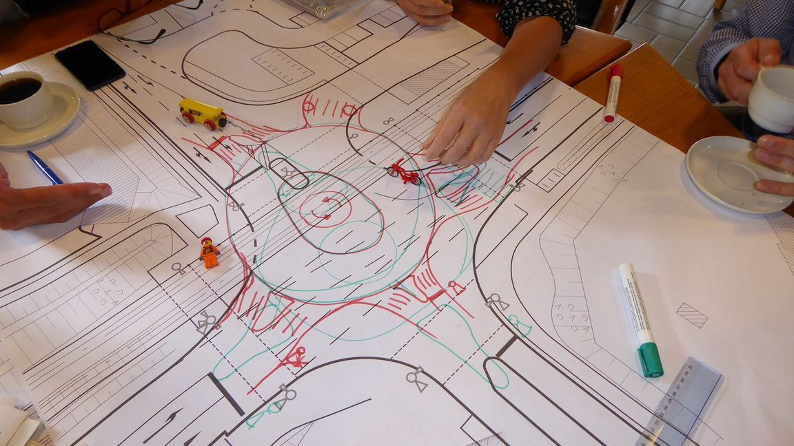 Hände von Teilnehmenden über einem Stadtplan mit Markierungen, Figuren und Spielzeugautos während eines Planungsworkshops.