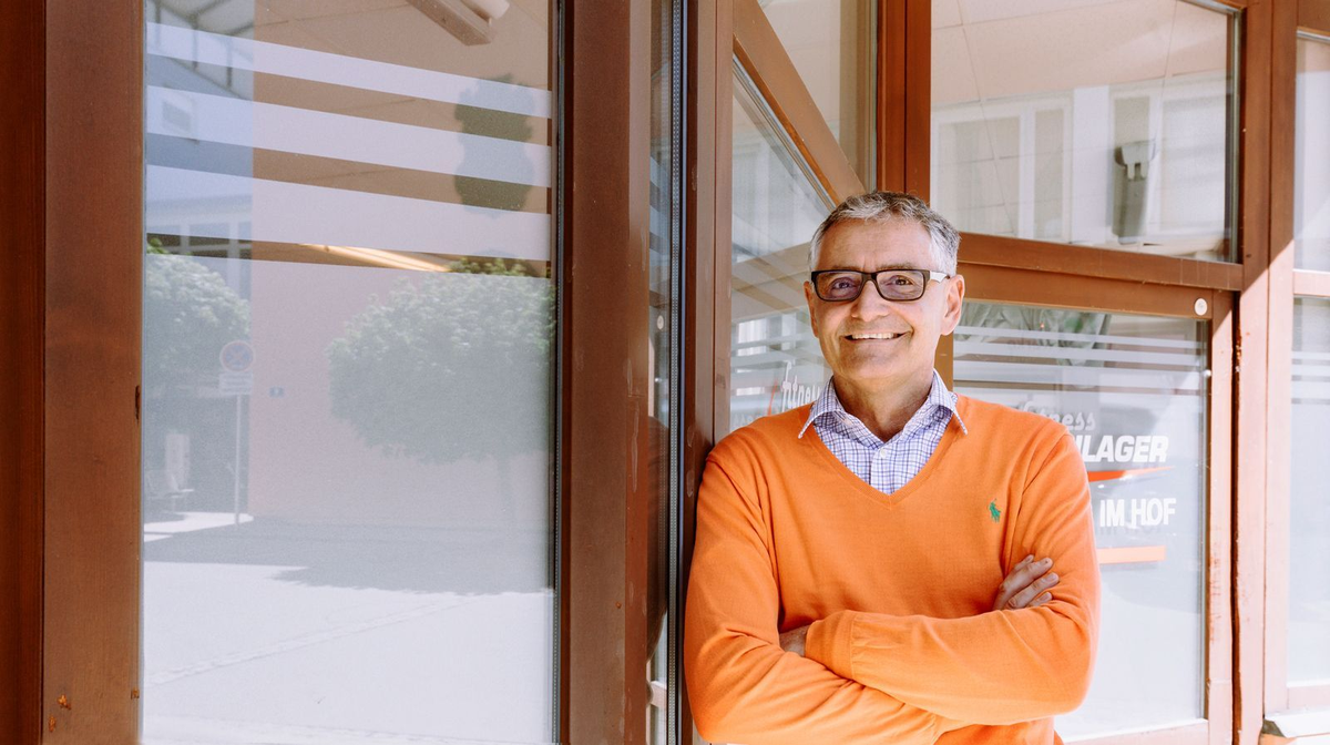 Person mit Brille und orangefarbenem Pullover steht lächelnd vor einer Glasfront in der Sonne.