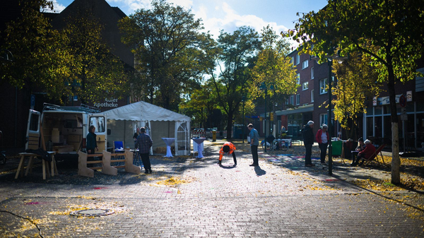 Menschen bereiten auf einer von Bäumen gesäumten Straße eine kleine Veranstaltung mit Zelt und Transporter vor, Herbstlaub liegt auf dem Boden.