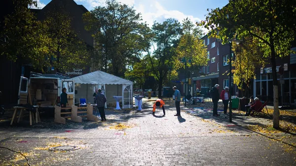Menschen bereiten auf einer von Bäumen gesäumten Straße eine kleine Veranstaltung mit Zelt und Transporter vor, Herbstlaub liegt auf dem Boden.