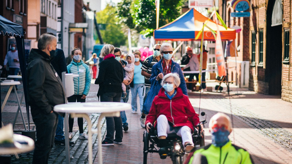 Menschen mit Masken in einer belebten Stadtstraße mit Ständen und buntem Zelt, vorne Person auf Liegerad.