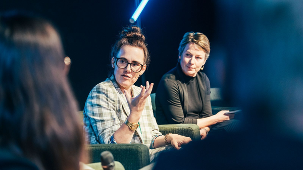 Zwei Frauen sitzen auf einer Bühne im Gespräch, eine spricht gestikulierend, die andere hört zu.