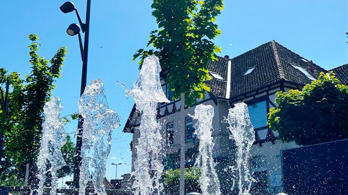 Sprudelnde Wasserfontänen eines Brunnens vor Bäumen und einem Haus mit Fachwerkdach bei strahlend blauem Himmel.