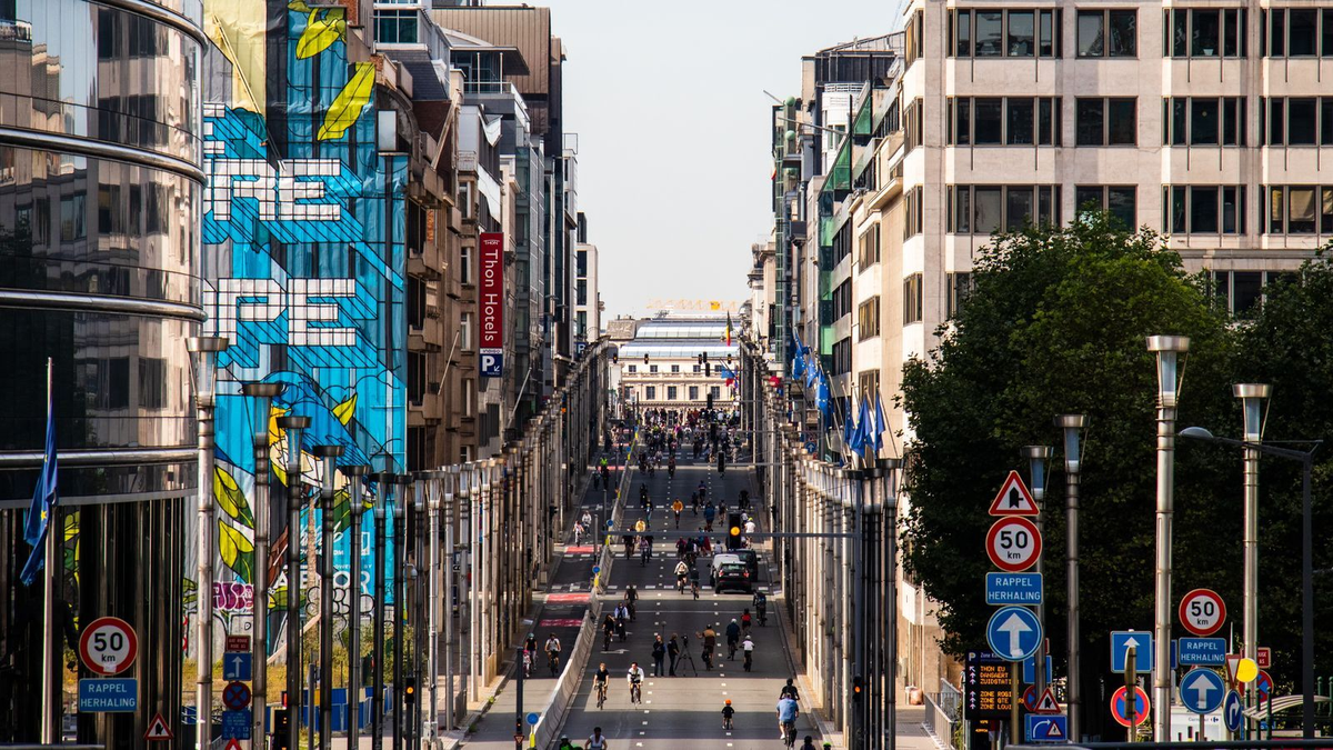 Städtische Straße mit vielen Radfahrenden zwischen modernen Gebäuden und EU-Flaggen, im Hintergrund ein historisches Gebäude.