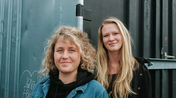 Zwei Frauen stehen nebeneinander vor einer blaugrauen Metallwand und blicken in die Kamera.