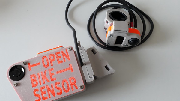 Zwei OpenBikeSensor-Geräte auf einem Tisch, graue 3D-gedruckte Gehäuse mit oranger Aufschrift und Kabeln zur Abstandsmessung im Radverkehr.