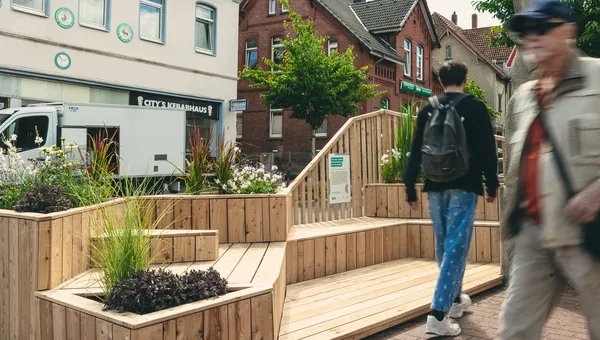 Ein hölzernes Parklet mit Sitzstufen und Pflanzkübeln auf einem früheren Stellplatz, zwei Passanten gehen vorbei.
