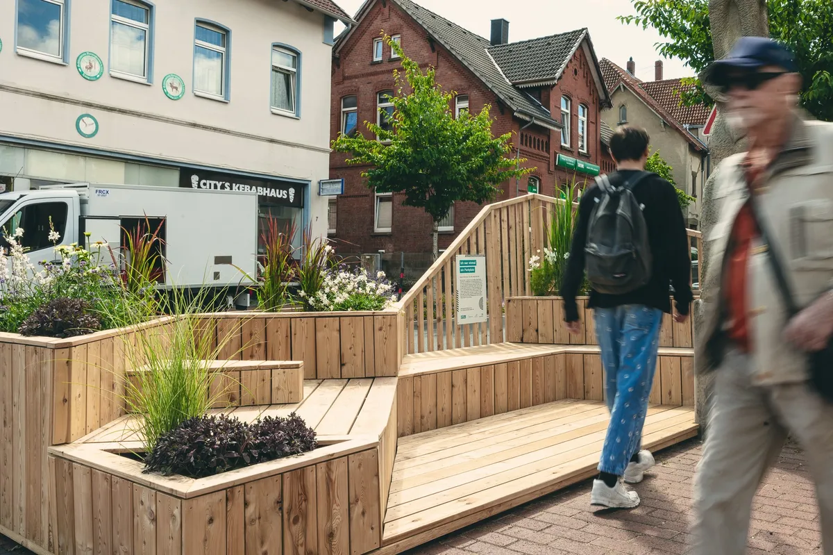 Ein hölzernes Parklet mit Sitzstufen und Pflanzkübeln auf einem früheren Stellplatz, zwei Passanten gehen vorbei.