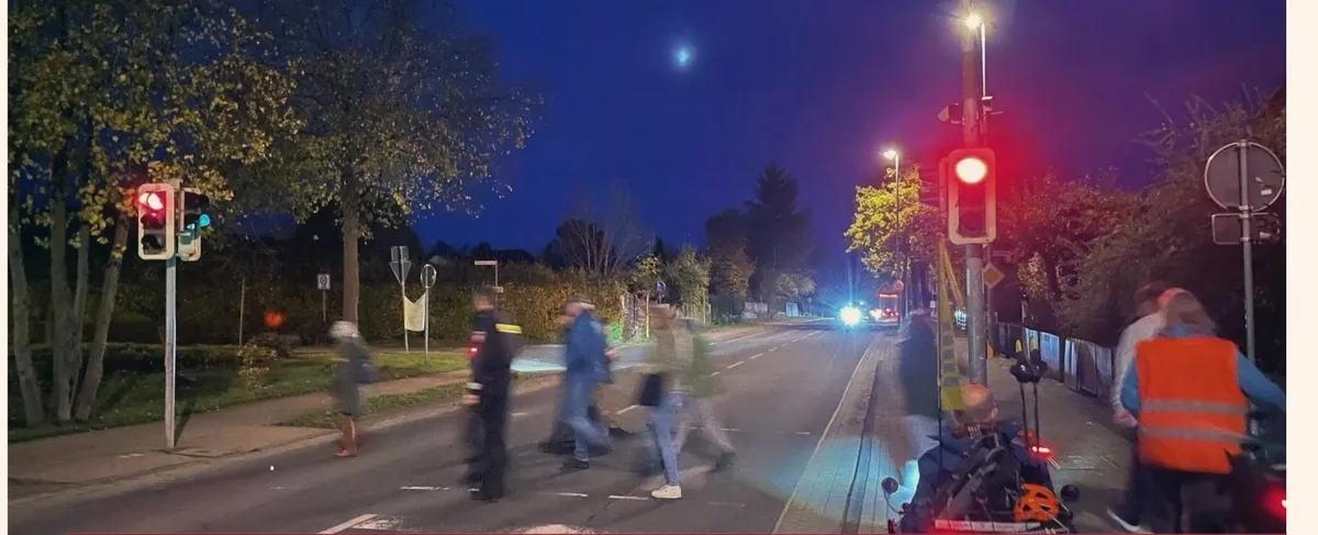 Menschen queren am Abend eine Hauptstraße an einer Fußgängerampel, die Rot zeigt.