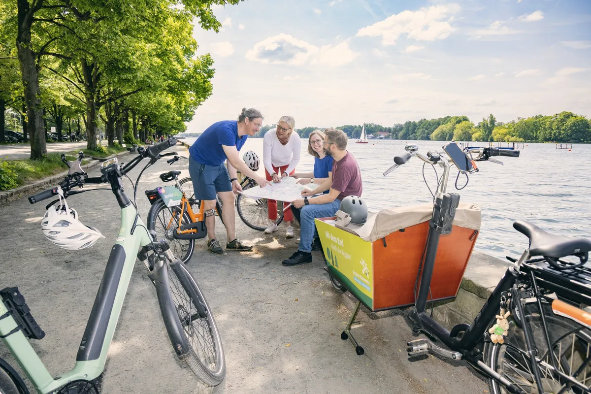 Vier Menschen mit Rädern und Lastenrad beugen sich am Ufer gemeinsam über eine Karte.
