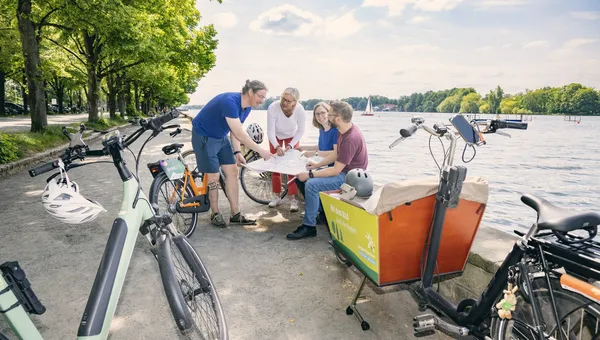 Vier Menschen mit Rädern und Lastenrad beugen sich am Ufer gemeinsam über eine Karte.
