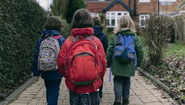 Drei Grundschulkinder mit Ranzen gehen nebeneinander einen Gehweg entlang.