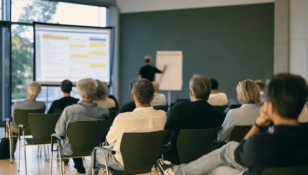 Blick aus der letzten Reihe in einen Seminarraum, vorn eine Präsentation und ein Flipchart.