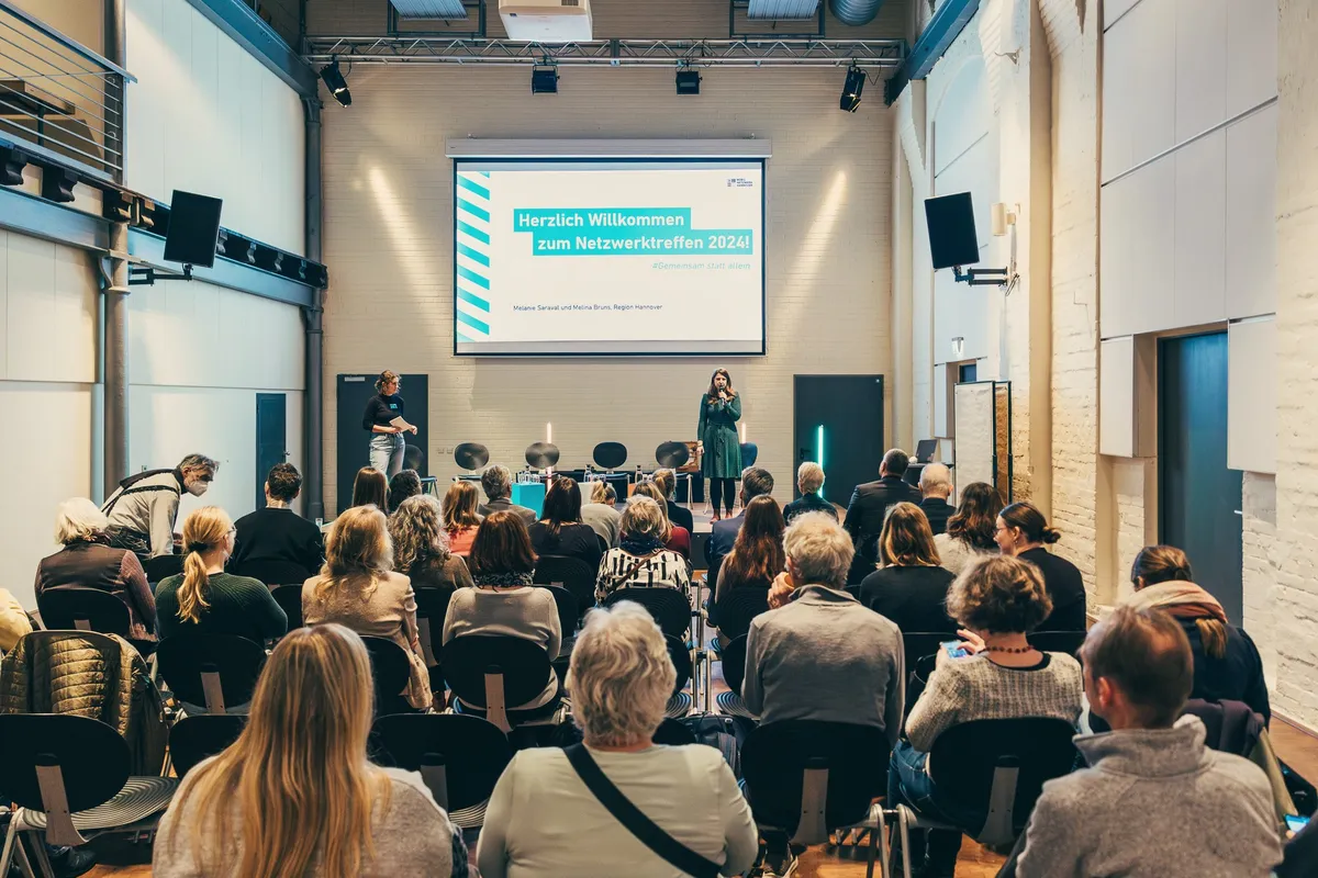 Publikum von hinten in einem vollen Saal, auf der Bühne die Begrüßungsfolie zum Netzwerktreffen.