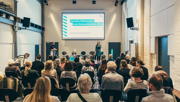 Publikum von hinten in einem vollen Saal, auf der Bühne die Begrüßungsfolie zum Netzwerktreffen.