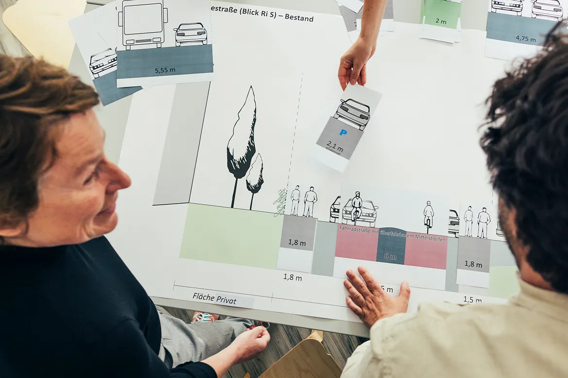 Drei Personen sitzen an einem Tisch und arbeiten gemeinsam an einem Straßenplan. Auf dem Plan sind Flächen für Gehwege, Radwege, Parken und Privatgrundstücke eingezeichnet. Mit Papierelementen visualisieren sie verschiedene Straßenbreiten und Verkehrsführ