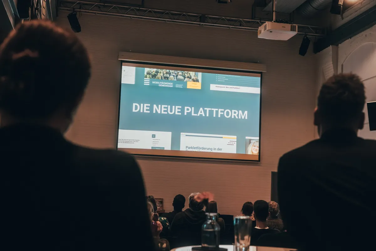 Leinwand zeigt die Folie „Die neue Plattform“, im Vordergrund sitzt das Publikum.