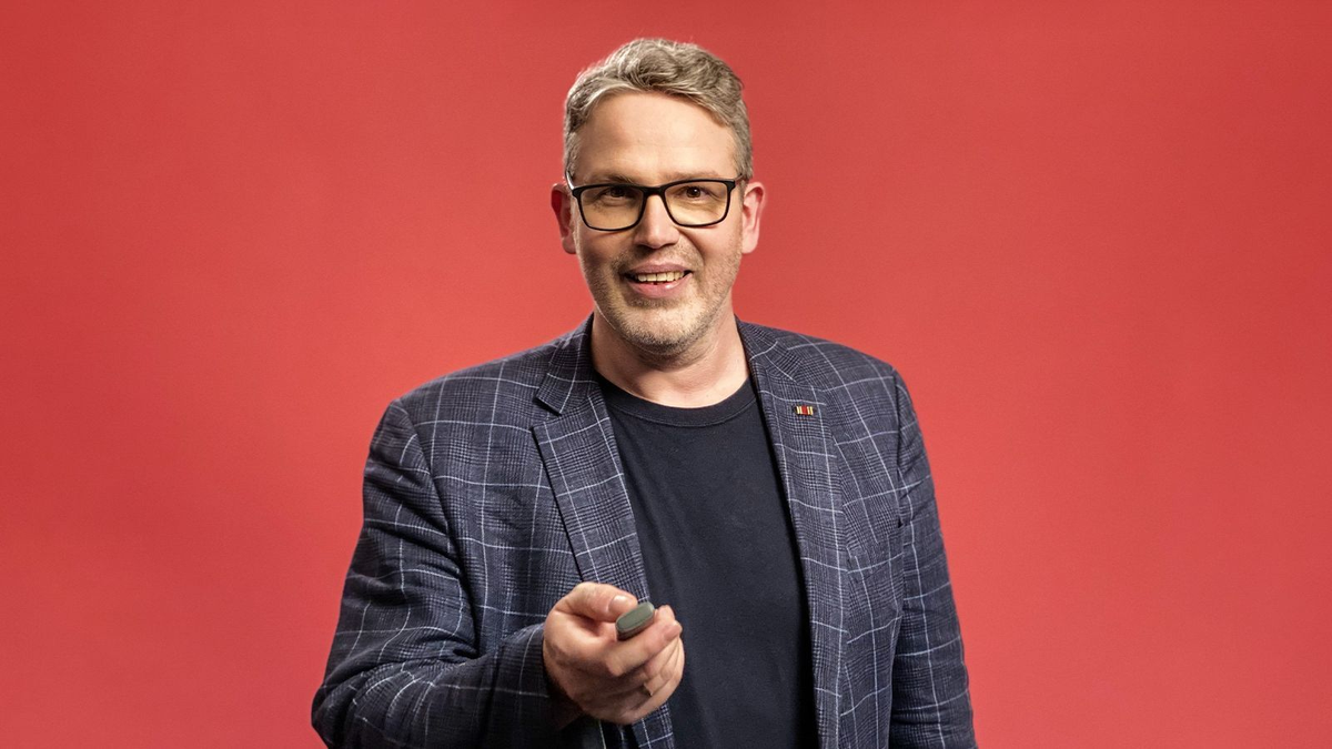 Lächelnde Person mit Brille und kariertem Sakko vor rotem Hintergrund, hält einen Presenter in der Hand.