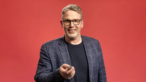 Lächelnde Person mit Brille und kariertem Sakko vor rotem Hintergrund, hält einen Presenter in der Hand.