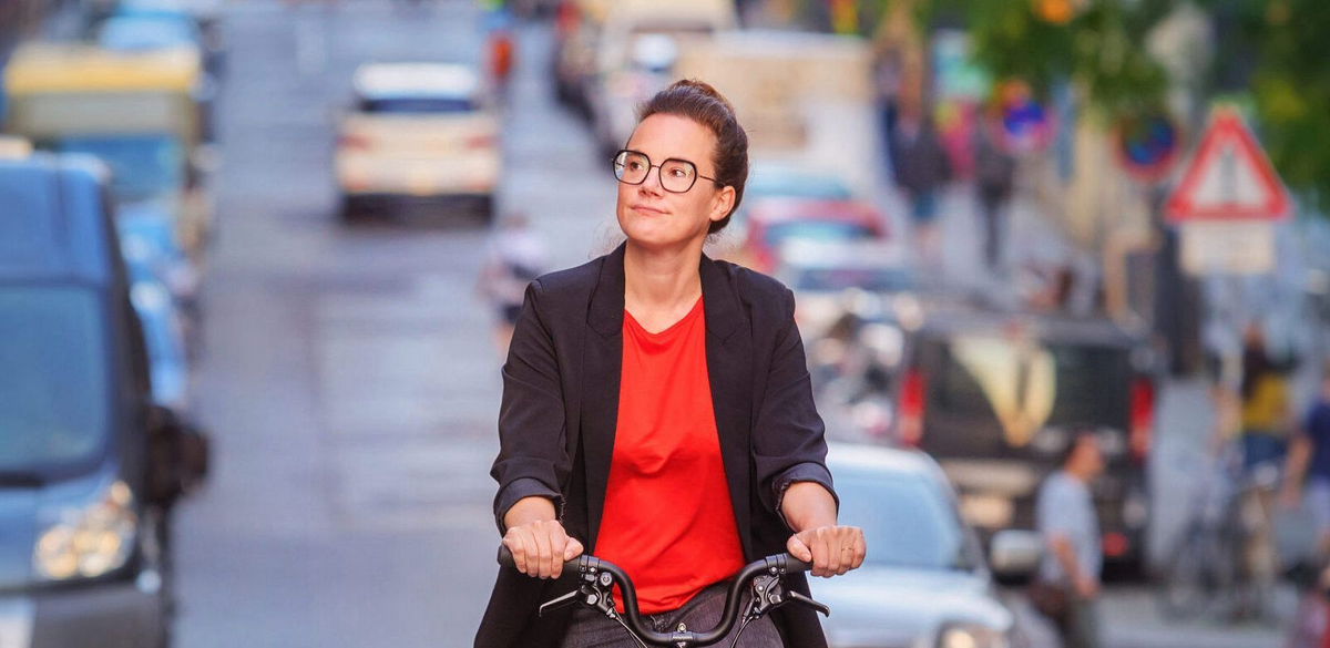 Katja Diehl mit Brille und rotem Shirt fährt mit dem Fahrrad durch eine Stadtstraße mit Autos und Fußgängern im Hintergrund.