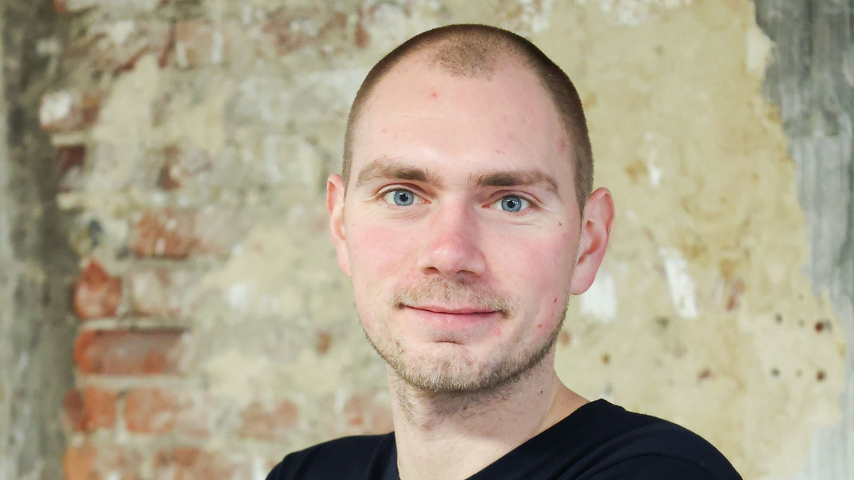 Porträt einer Person vor einer Wand aus hellem, freigelegtem Mauerwerk mit leichtem Lächeln in die Kamera blickend.