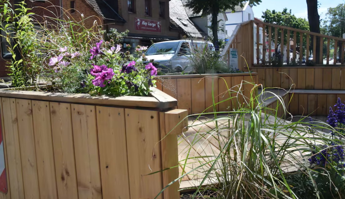 Holz-Parklet mit bepflanzten Kästen und Sitzbereich an einer sonnigen Stadtstraße.