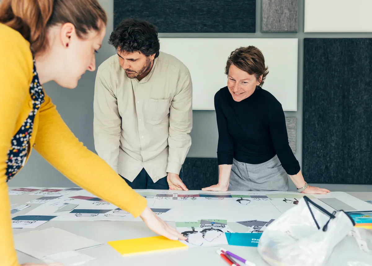 Drei Personen stehen an einem Tisch und arbeiten gemeinsam an einem großformatigen Plan.