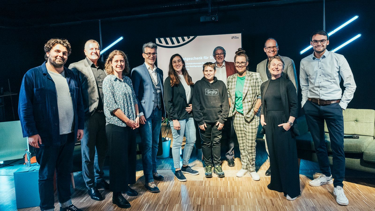 Gruppenfoto von zwölf Personen auf einer Bühne vor einer Leinwand bei einer Veranstaltung, freundlich in die Kamera blickend.