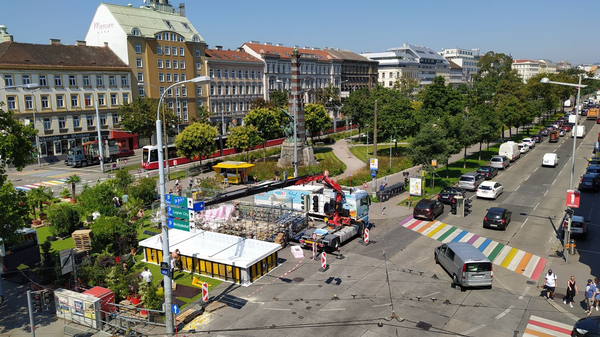Stadtansicht von Wien mit bunter Kreuzung, historischen Gebäuden, Parkanlage und Verkehr bei sonnigem Wetter.