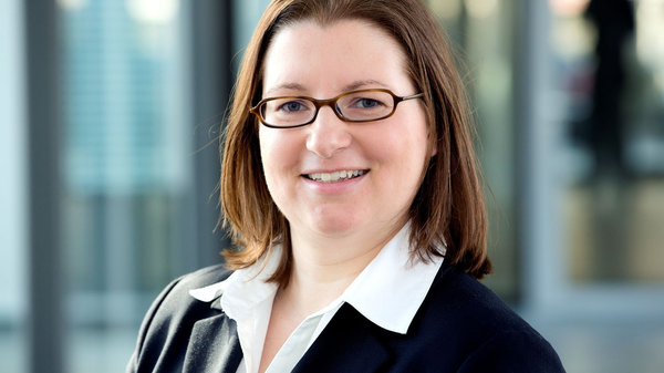 Porträt einer Frau mit Brille, weißem Hemd und dunklem Sakko vor Bürohintergrund.