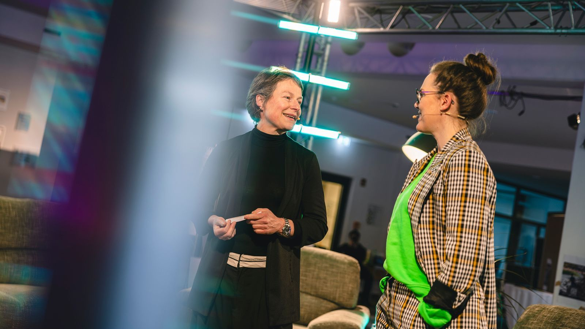 Zwei Frauen sprechen auf einer Bühne miteinander, beide mit Mikrofonen, im Hintergrund leuchten bunte Lichter.