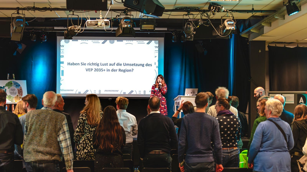 Frau spricht auf Bühne vor Publikum, hinter ihr steht auf Leinwand „Haben Sie richtig Lust auf die Umsetzung des VEP 2035+ in der Region?“.