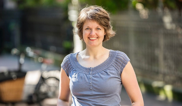 Porträt einer lächelnden Frau im Freien mit grauem Oberteil und unscharfem Hintergrund mit Fahrrad und Zaun.