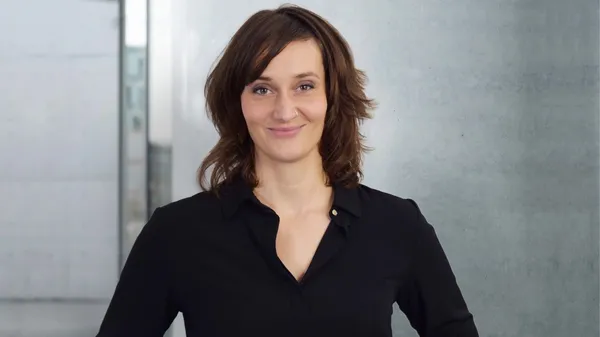 Porträt einer Frau mit braunen Haaren und schwarzer Bluse vor modernem, hellgrauem Hintergrund.