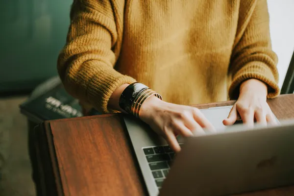 Eine Person sitzt an einem Tisch und tippt während eines Webinars auf einem Laptop. Sie trägt einen gelben Strickpullover und mehrere Armreifen. Neben ihr liegt ein Notizbuch, das Bild ist warm beleuchtet.