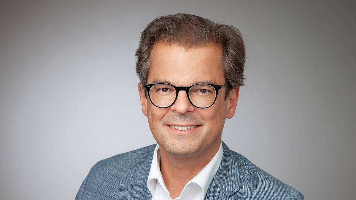 Porträt eines Mannes mit Brille, blauem Sakko und weißem Hemd vor grauem Hintergrund, freundlich lächelnd.